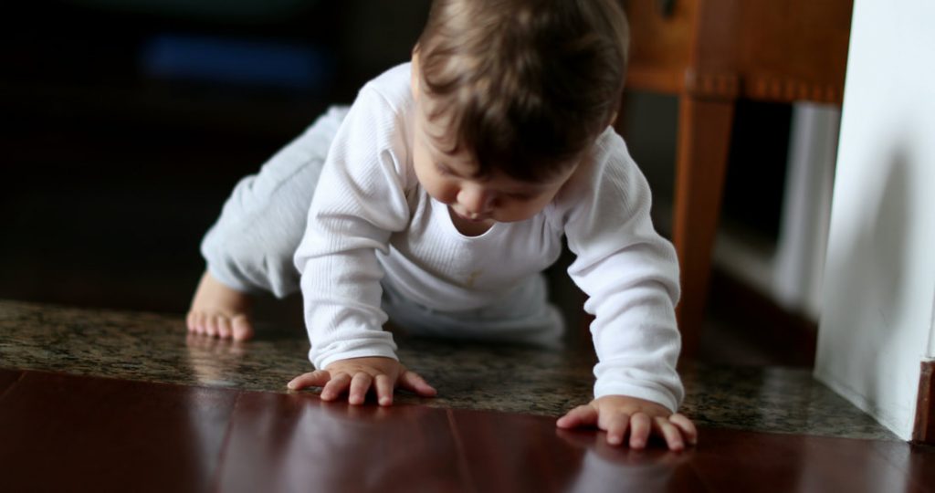 Bébé en body blanc rampant avec curiosité sur un sol en bois lustré, explorant son environnement, représentatif des étapes de développement de la petite enfance chez GraineSante