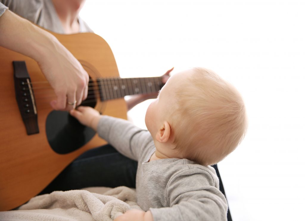 Bébé en émerveillement touchant une guitare acoustique jouée par un adulte, capturant un moment d'éveil musical précoce sur fond blanc lumineux.